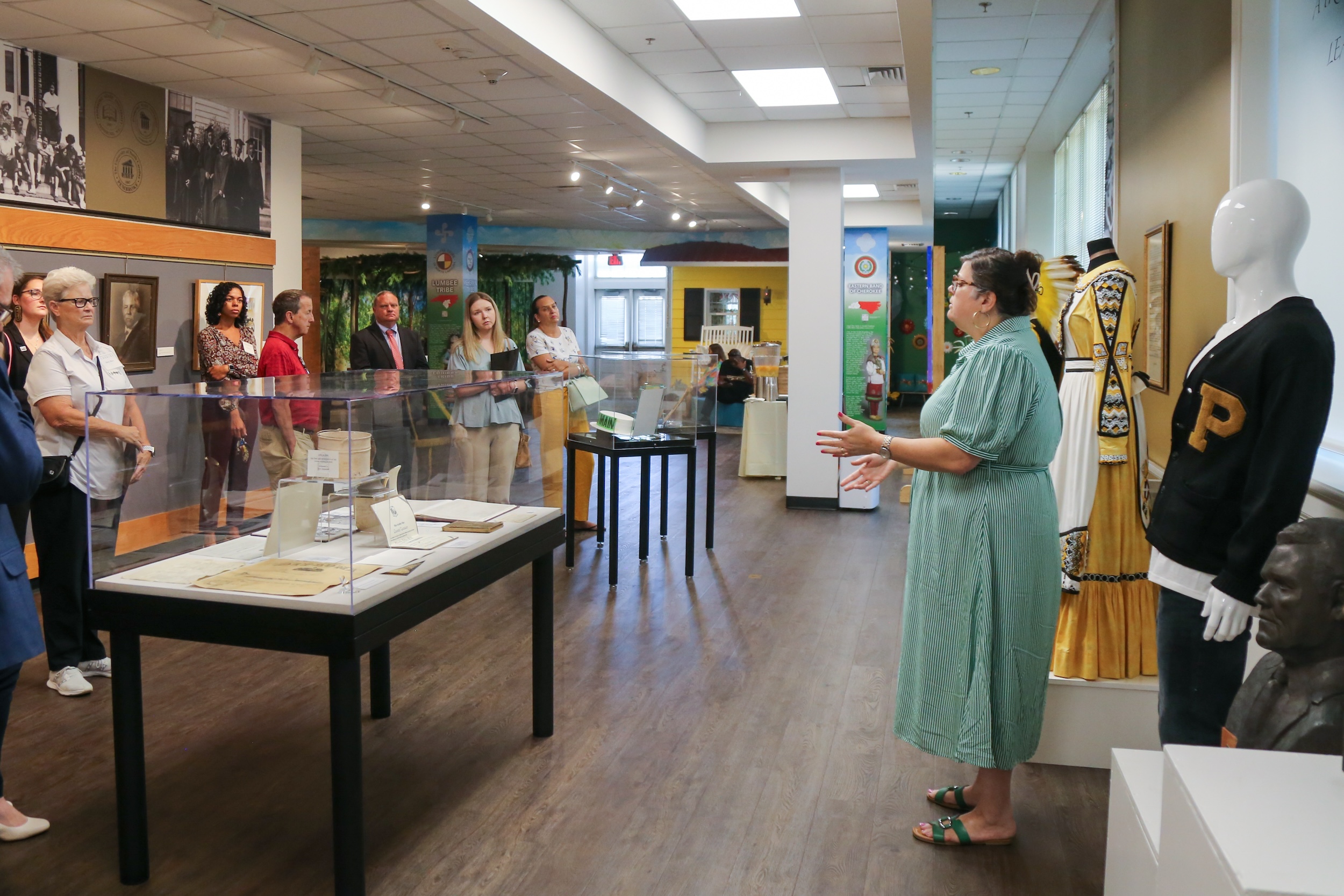 A guide leads a group tour through the Museum of the Southeast American Indian, with historical documents in display cases, cultural regalia on mannequins, and historic photographs lining the walls.