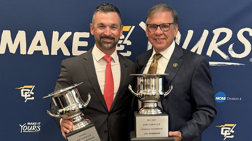 UNCP Athletics Director Dick Christy, left, with Chancellor Robin Gary Cummings, right, both holding large silver trophies.