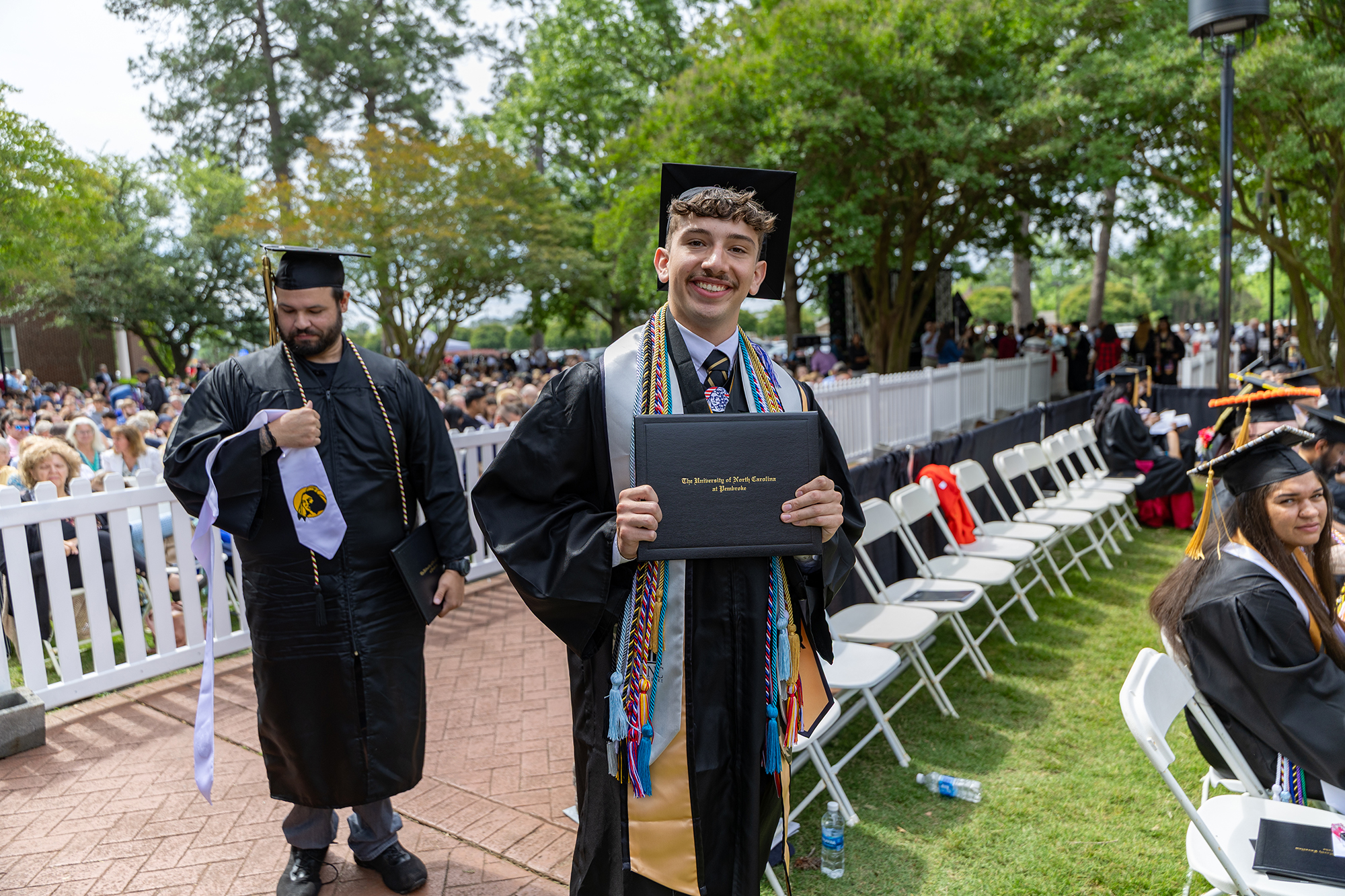 UNCP student John Squire holds his diploma and smiles while dressed in his cap and gown.