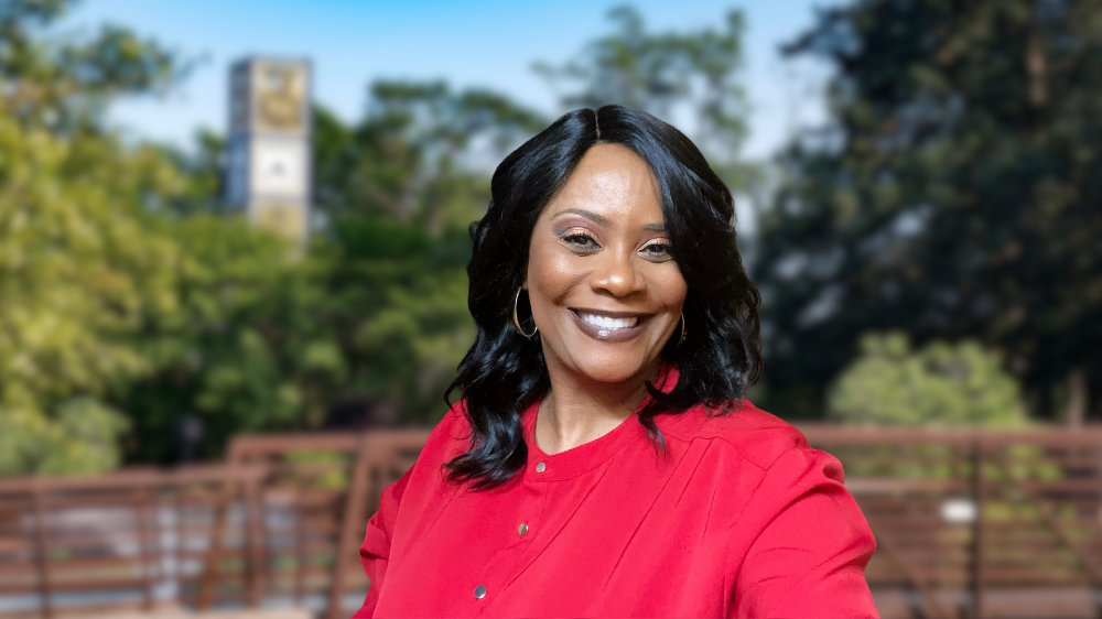Michelle McNeil smiles for a portrait while wearing a red blouse. She is pictured outdoors with a blurred campus background, including trees, a walkway and a campus tower in the distance.