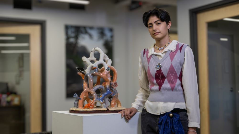 Mia Baca stands beside their coral sculpture in an indoor campus setting at UNC Pembroke. The piece features twisting, branch-like coral forms in soft white, peach and blue tones, visually reflecting the beauty and fragility of ocean life.