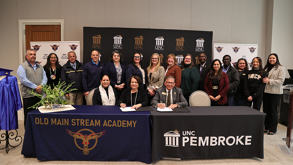 UNC Pembroke Chancellor Robin Gary Cummings and Dr. Rose Marie Lowry-Townsend, chair of Old Main STREAM Academy, sign a dual enrollment agreement while staff from both institutions look on.