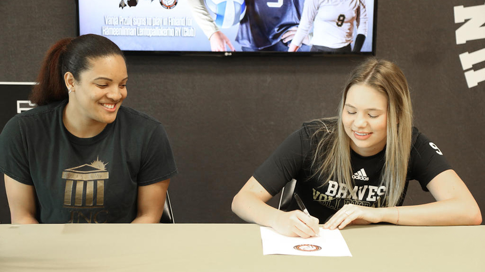 UNC Pembroke volleyball player signs professional contract while seated beside a smiling coach, with a screen in the background announcing the signing.