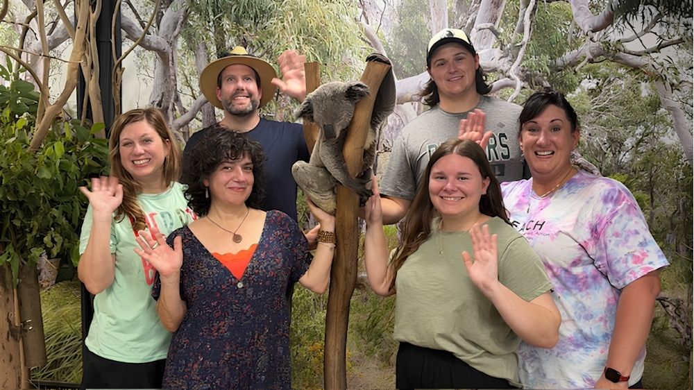 A group of six UNC Pembroke REACH participants pose and wave beside a koala exhibit during a trip to Australia