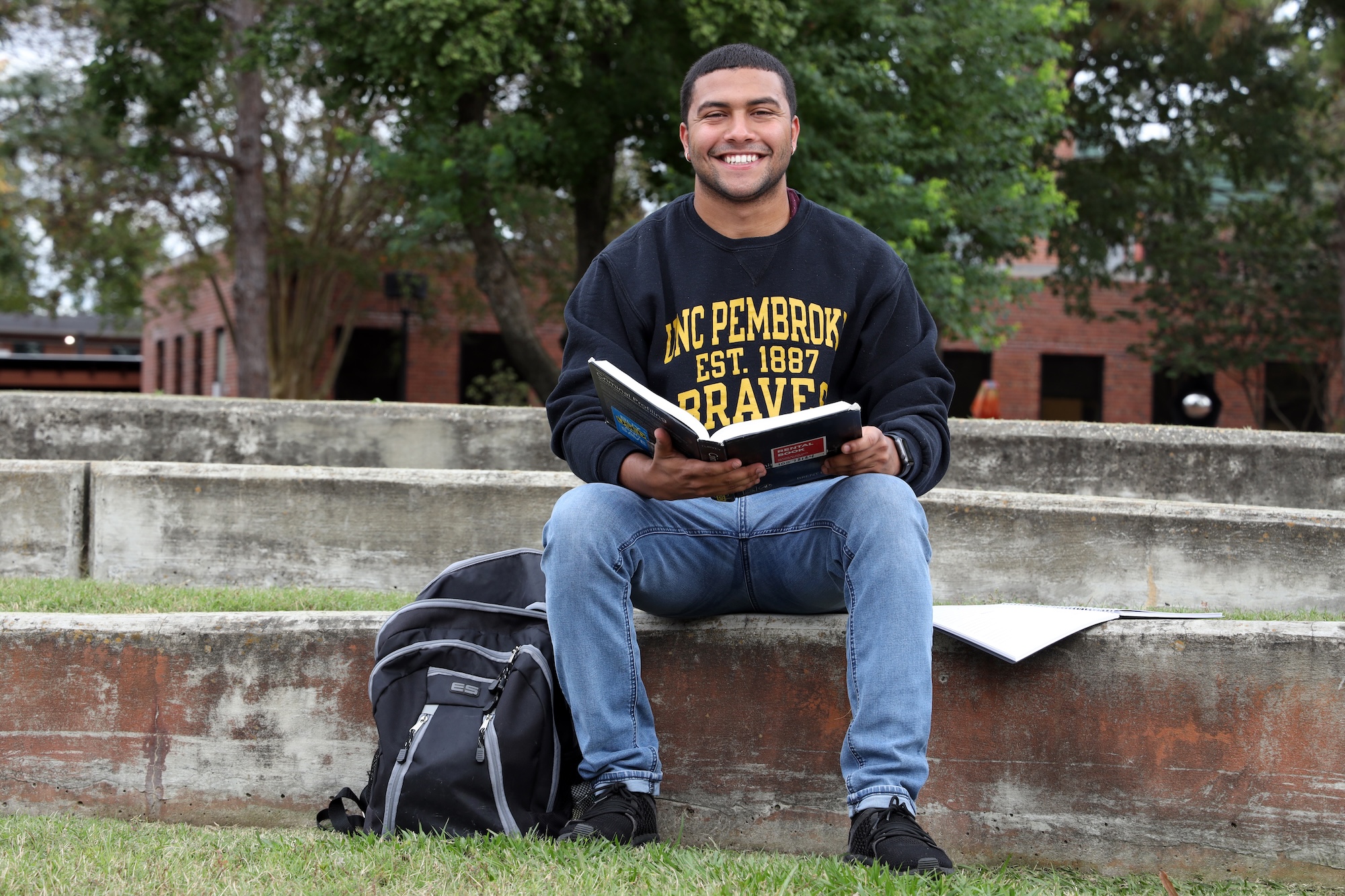 UNCP Criminal Justice graduate Cameron Richardson sits on campus reading a textbook while wearing a UNC Pembroke Braves sweatshirt, representing the student experience in the Criminal Justice program.