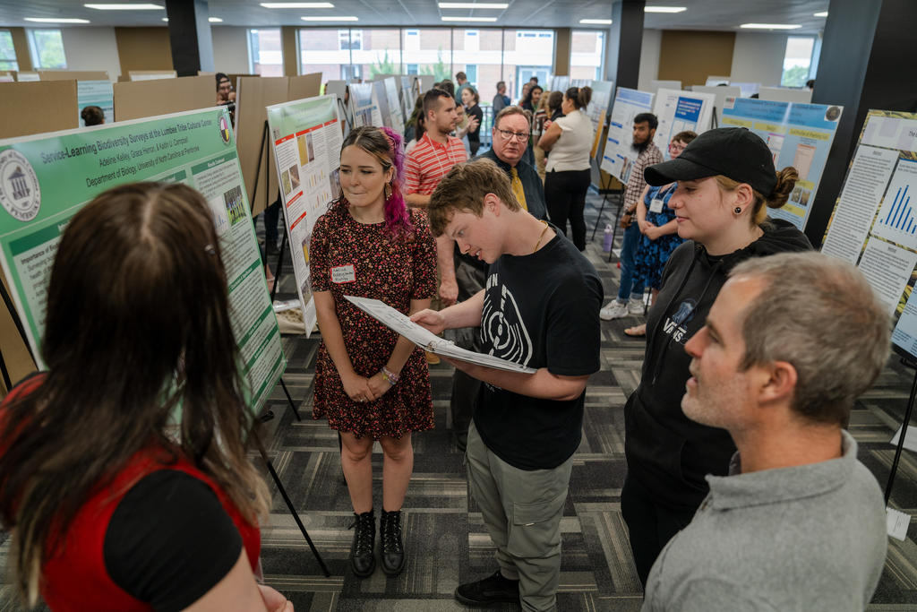 Students and faculty engage with a presenter during a poster session at UNC Pembroke’s research symposium, surrounded by rows of academic displays.