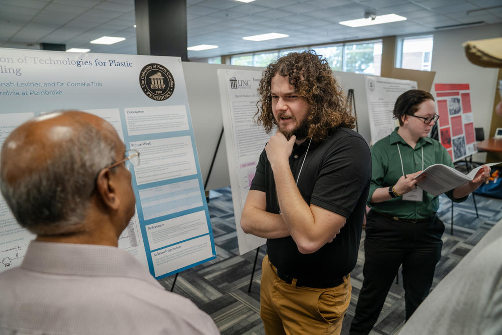 Students and faculty engage with a presenter during a poster session at UNC Pembroke’s research symposium, surrounded by rows of academic displays.