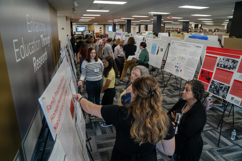 Students and faculty engage with a presenter during a poster session at UNC Pembroke’s research symposium, surrounded by rows of academic displays.