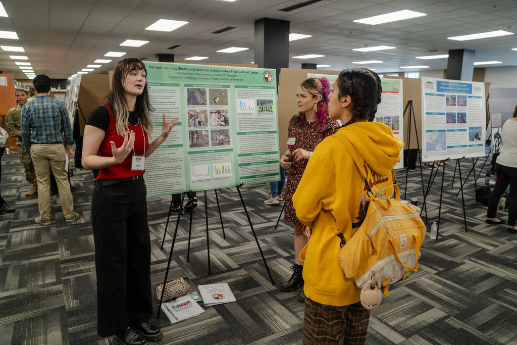 Students and faculty engage with a presenter during a poster session at UNC Pembroke’s research symposium, surrounded by rows of academic displays.