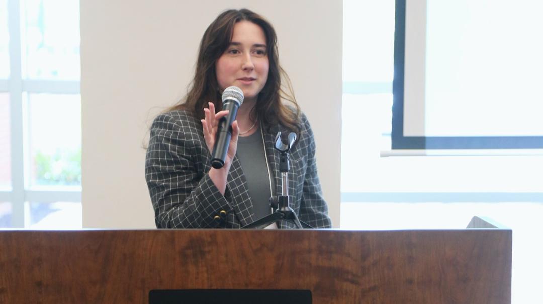 Female student at a podium, holding a microphone and presenting her research at a live talk in the library at the undergraduate research symposium at UNC Pembroke.
