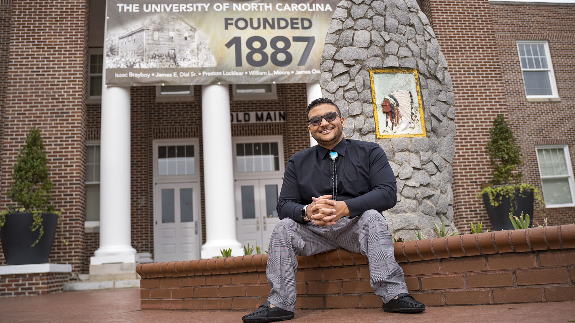 Benjamin Clark sitting and smiling in front of the Old Main building at UNC Pembroke, next to a stone monument with a painted image of a Native American chief.