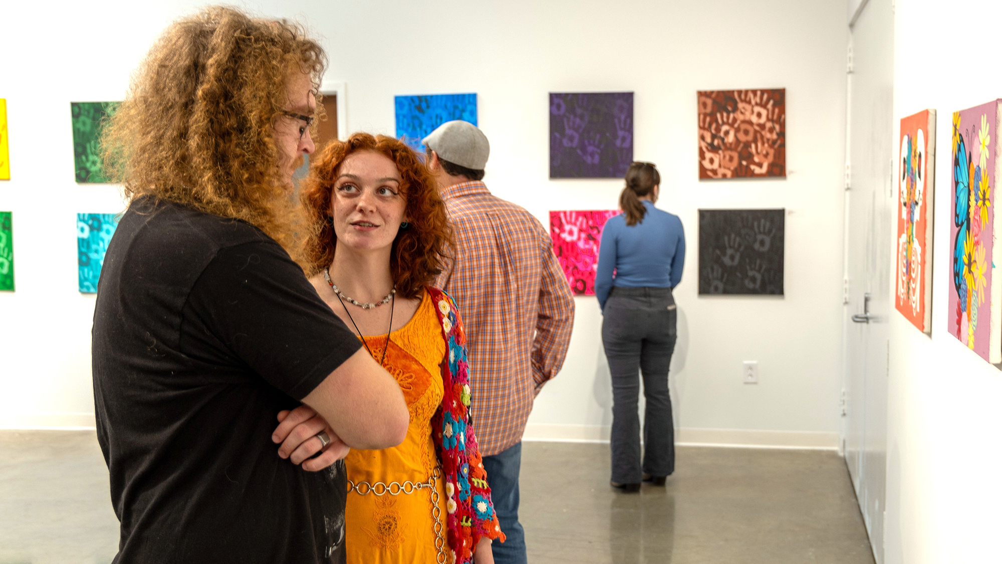 Two college students talk while admiring paintings mounted on the wall in front of them. Other students and public stand in the background looking at other works on other walls.