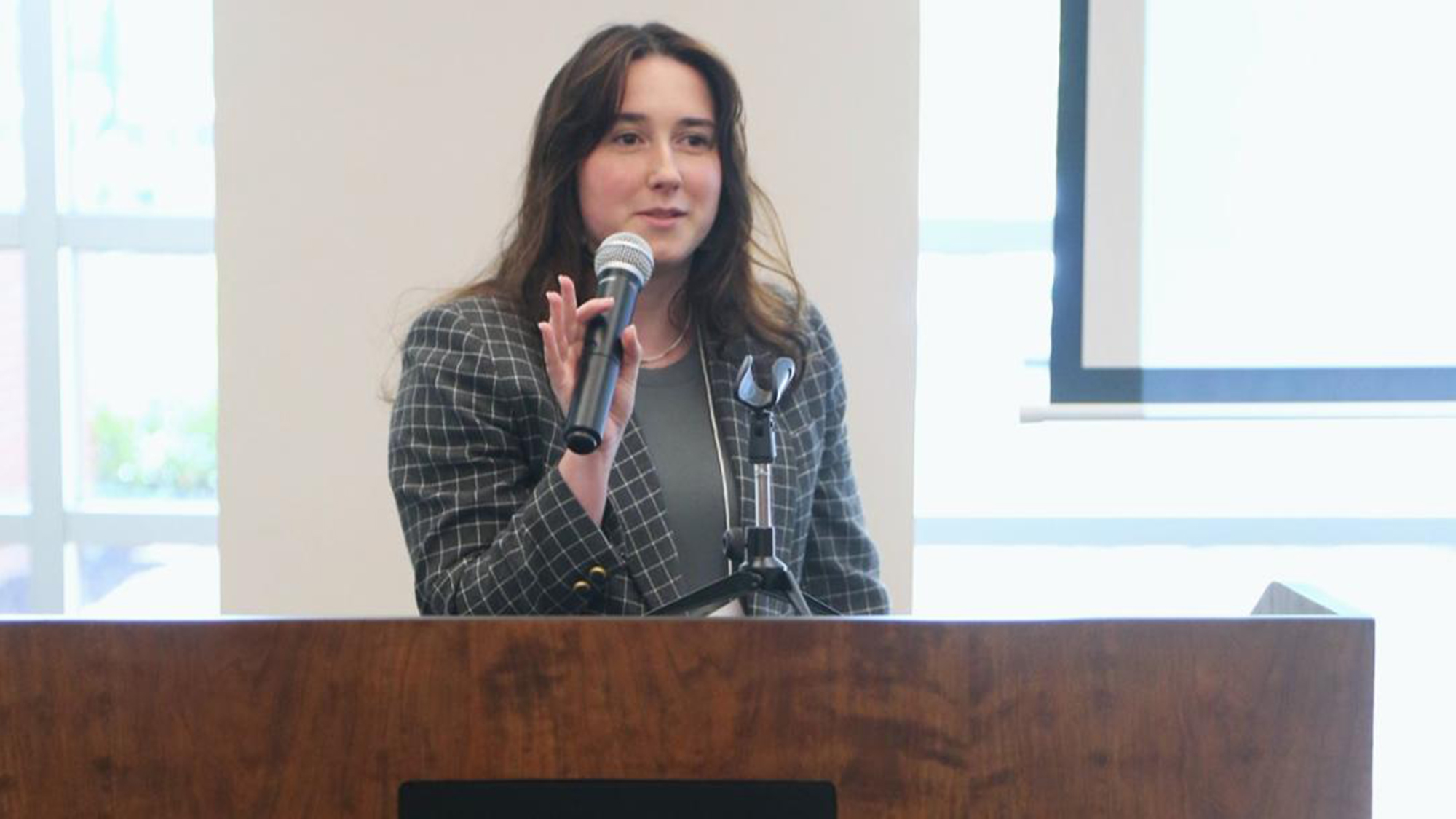 Female student at a podium, holding a microphone and presenting her research at a live talk in the library at the undergraduate research symposium at UNC Pembroke.