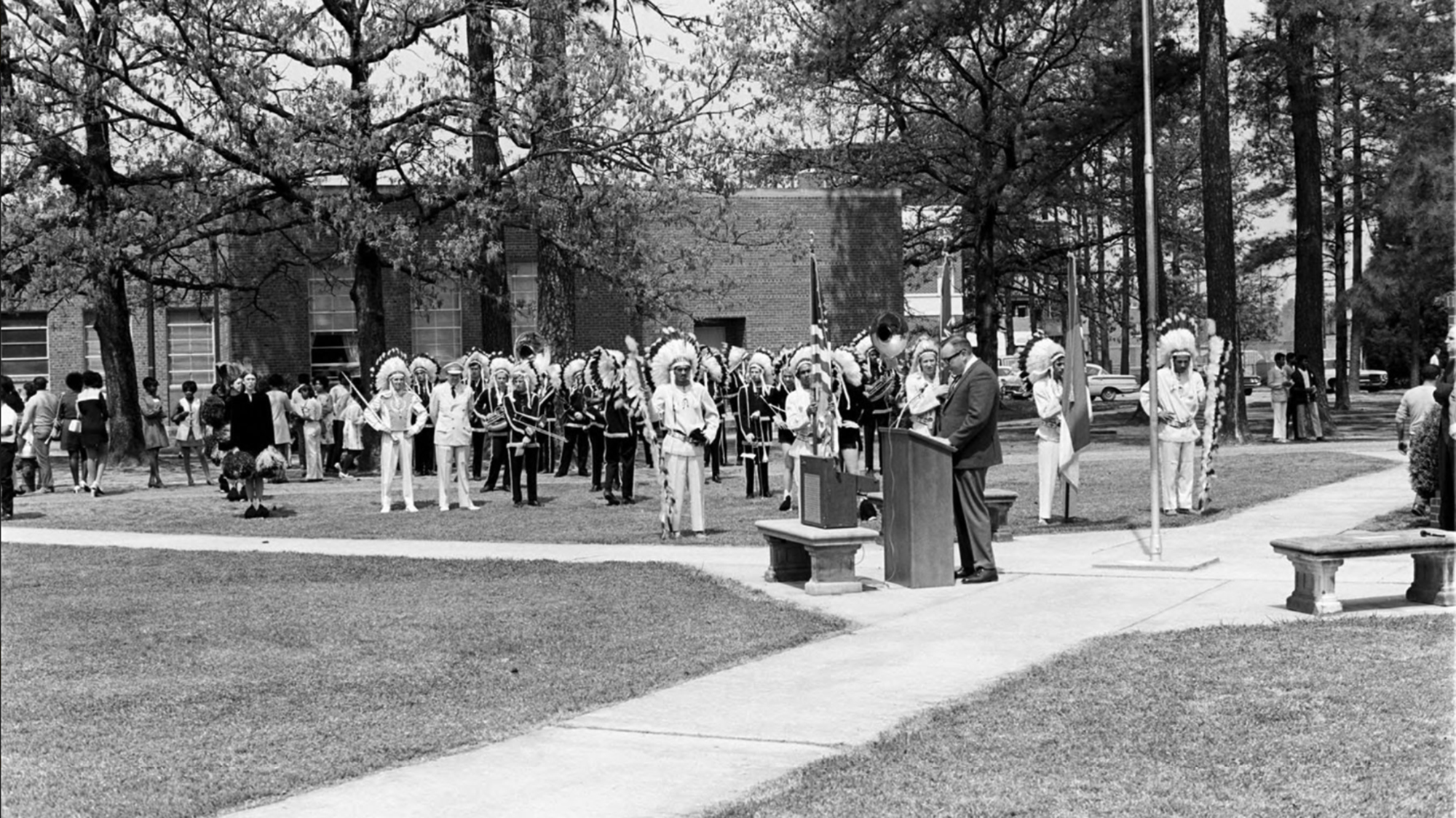 Chancellor speaking to crowd with marching band in ranks to his side at the intersection of two sidewalks