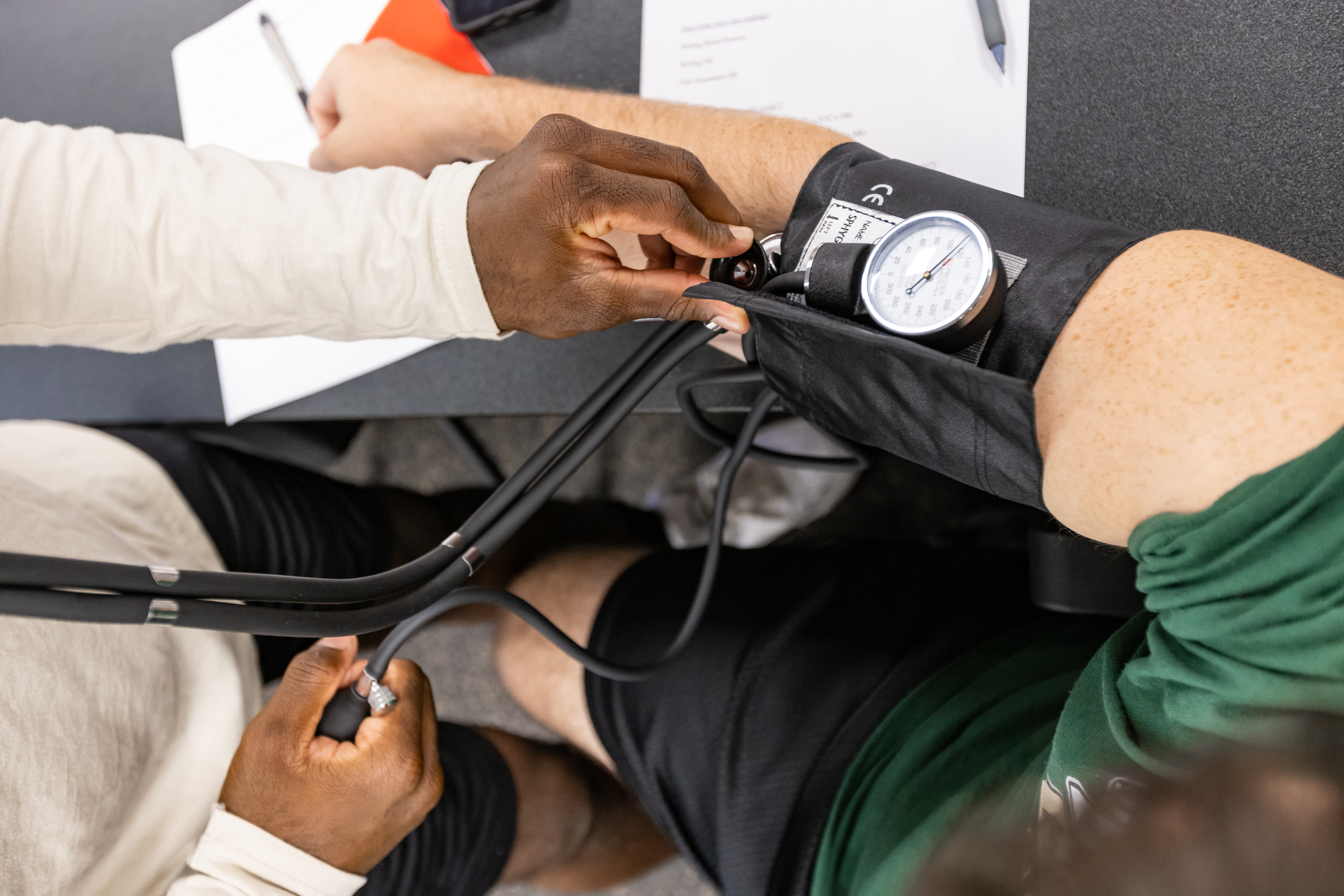 A person measuring another person's blood pressure using a sphygmomanometer. The person being measured is seated, and the cuff of the sphygmomanometer is wrapped around their upper arm. Another individual is operating the device, adjusting the valve and reading the gauge. There are papers and a pen on the table in front of them.