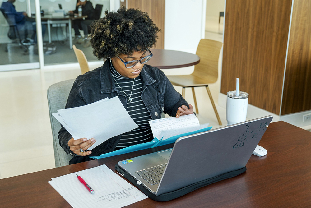 female student flipping through pages in a notebook while sitting at a desk with open laptop