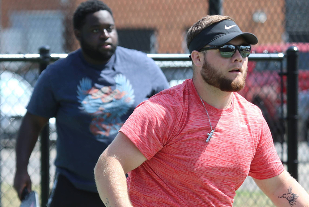 Two teammates playing pickleball on an outdoor court. The person in the foreground is wearing a red shirt and a black visor. The person in the background is wearing a dark blue shirt