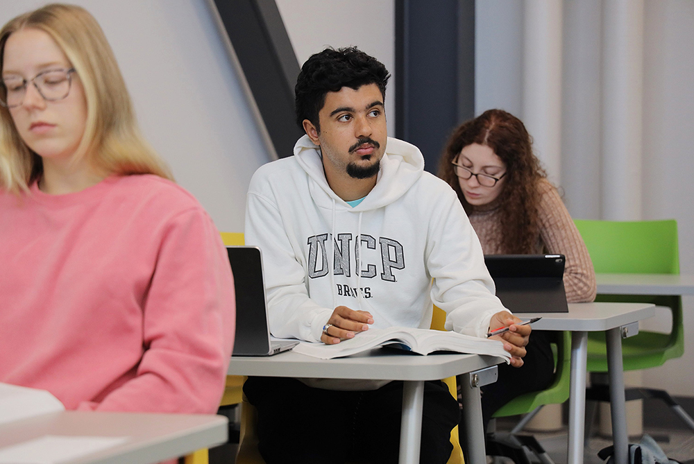 Male student in a white UNC Pembroke hoodie at a desk in a colorful classroom. Female stuents sit behind and in front of him. He's looking thoughtfully up and to the front of the classroom. The women are both intent on the work in front of them on their desks.