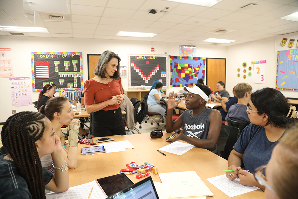 A classroom setting with several students seated at tables, engaging in group activities or discussions. A teacher is standing and interacting with one of the groups. The classroom walls are decorated with colorful educational posters and charts.