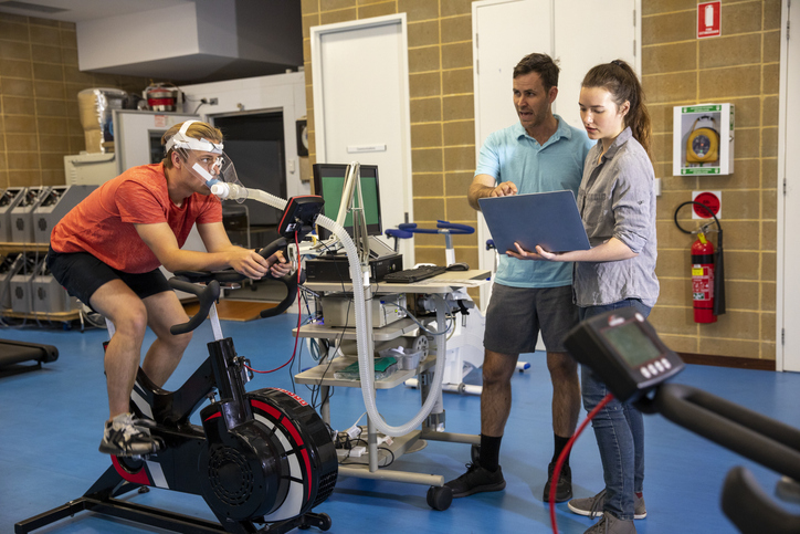 A person riding an exercise bike while wearing a mask connected to various monitoring equipment. Two other individuals are standing nearby, one holding a laptop and the other pointing at the screen. The setting appears to be a laboratory or medical facility with various pieces of equipment and storage units in the background.