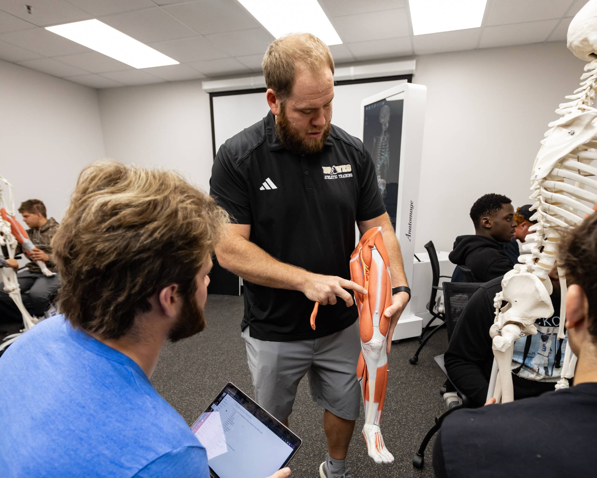 A classroom setting where a person, likely an instructor, is holding and pointing to a model of the human leg muscles. The instructor is explaining something to students who are seated around with laptops and notebooks. There is also a skeleton model visible in the background.