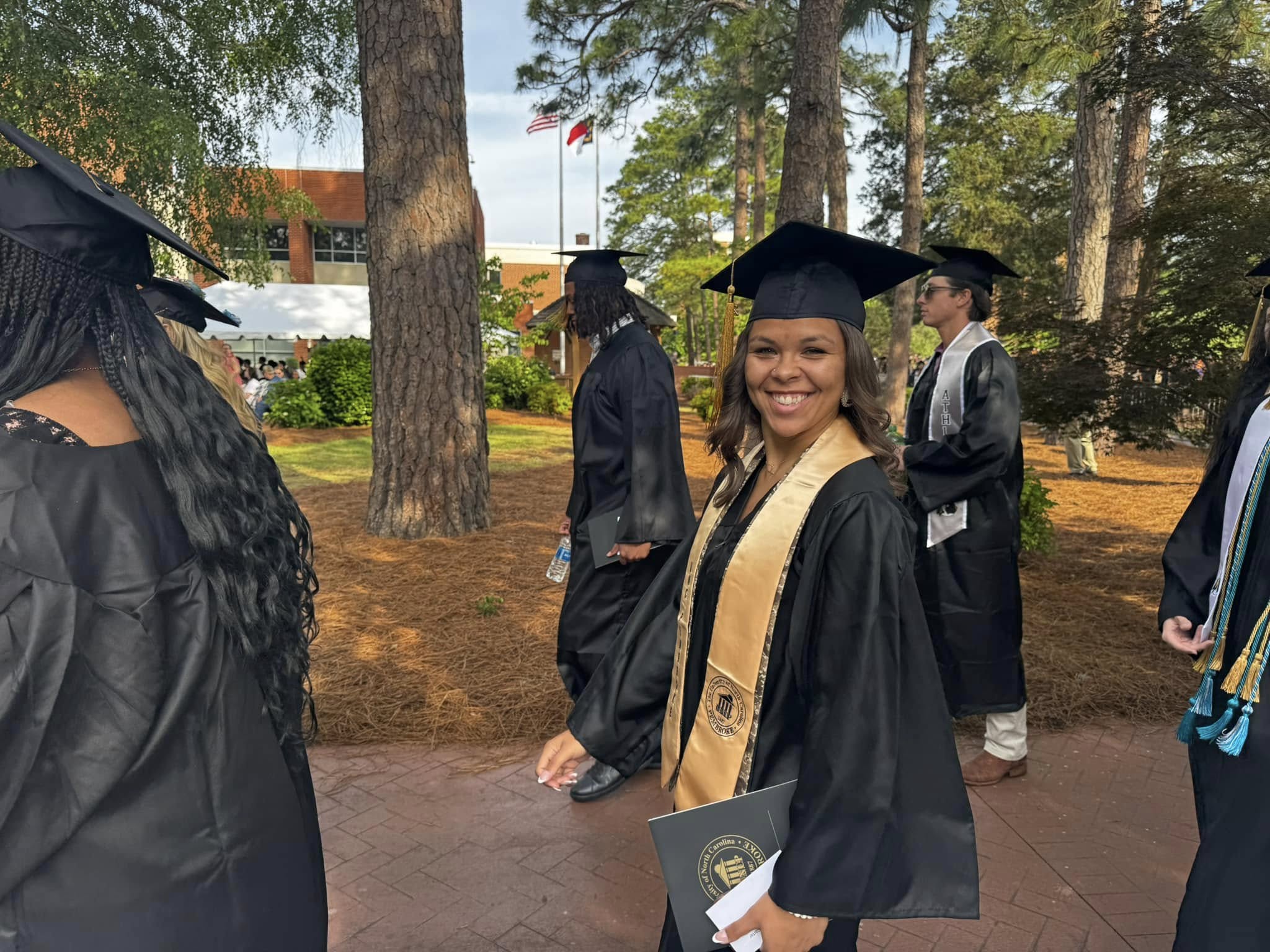 Graduates in caps and gowns walking on a brick pathway outdoors, surrounded by trees. Some hold diplomas, with flags, a building, and greenery visible in the background.