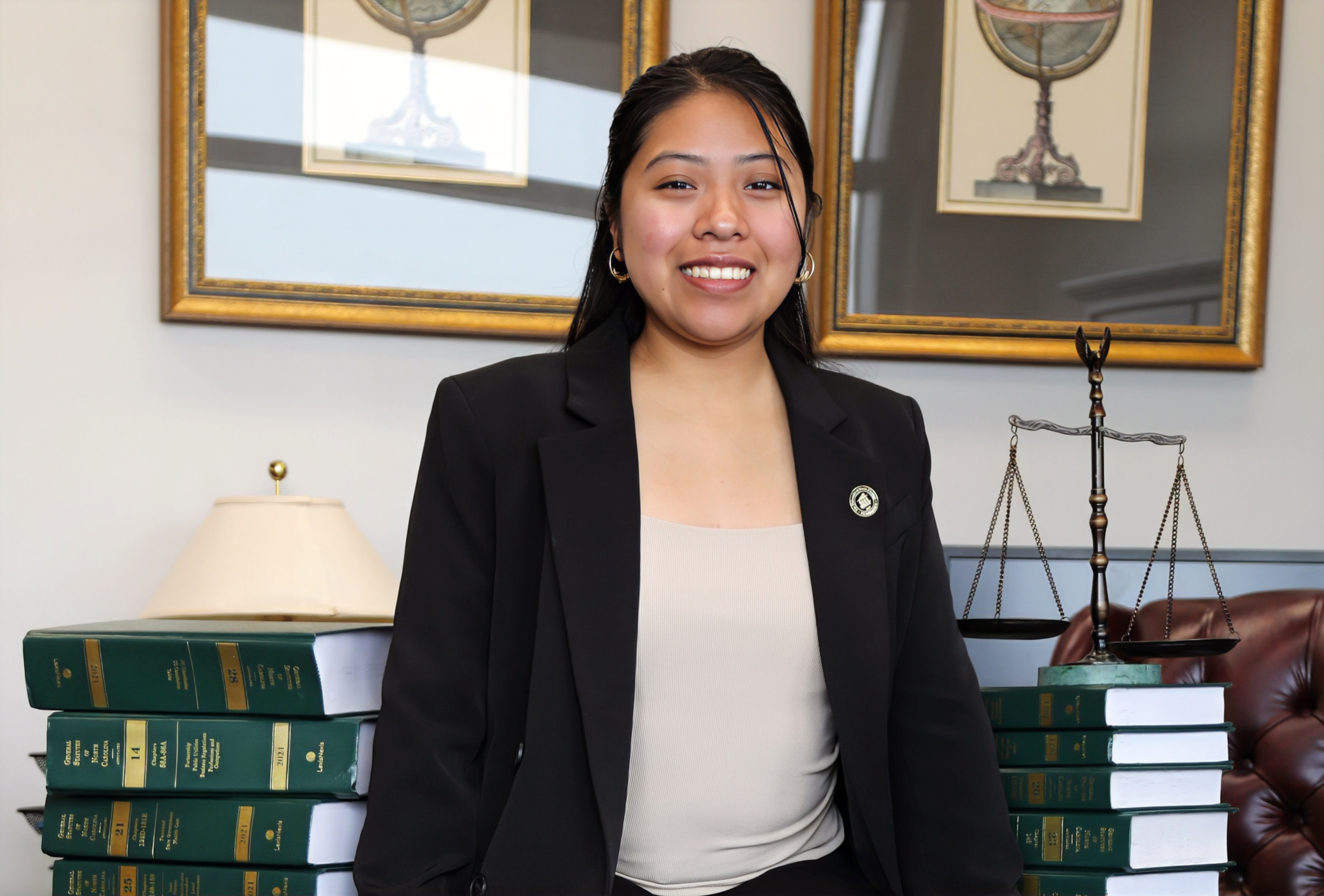 Magally Ortiz-Rojas, Political Science major, seated on a desk between two stacks of legal books with a set of scales on the stack to her left.