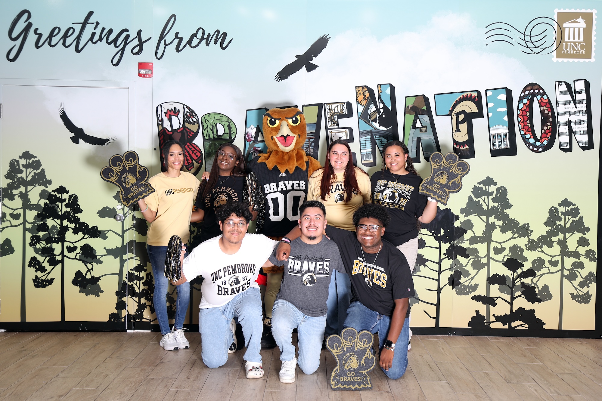 UNCP orientation leaders pose with the Braves mascot in front of a "Greetings from BraveNation" mural, wearing UNCP branded apparel.