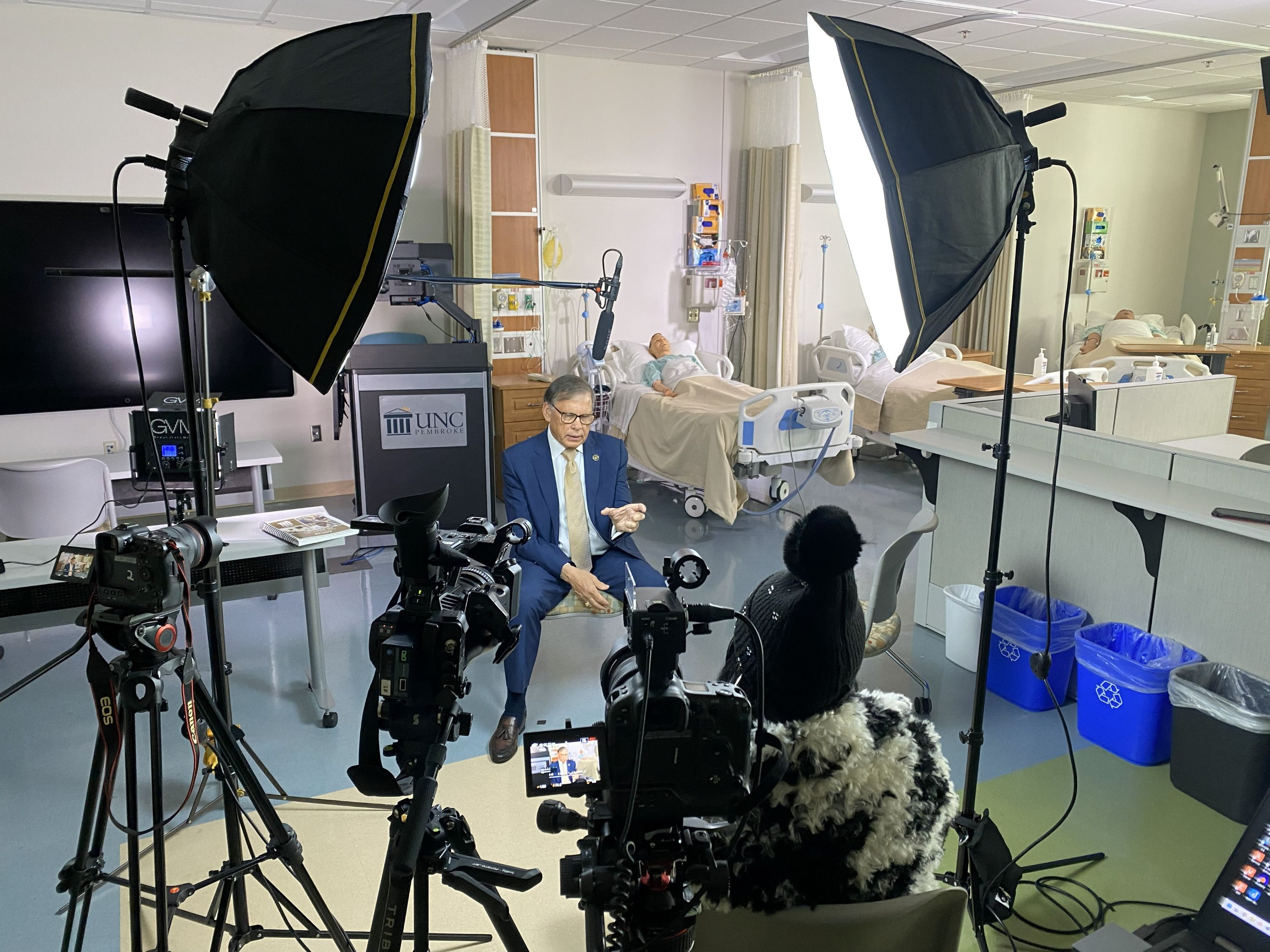 Behind-the-scenes setup for a Chancellor Robin G. Cummings media interview in the UNCP nursing simulation lab, with lighting and camera equipment visible.