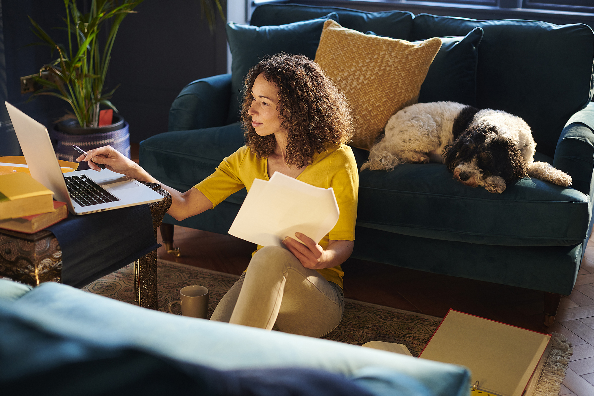 woman sitting on floor of her living room with dog behind her and she is working on her laptop